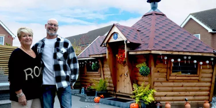 Mark and Anne stood in front of their sloping walled BBQ cabin with a side extension and red roof.