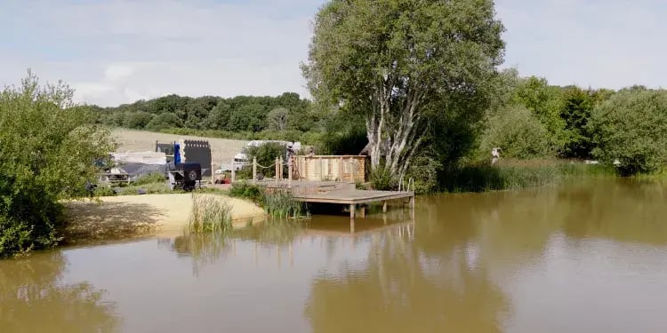 An Arctic Cabin being built next to a lake. The cabin has the walls standing in their place. To the left there are various pannels and materials for bulding the cabin. On the riverbank there is a timber pontoon.