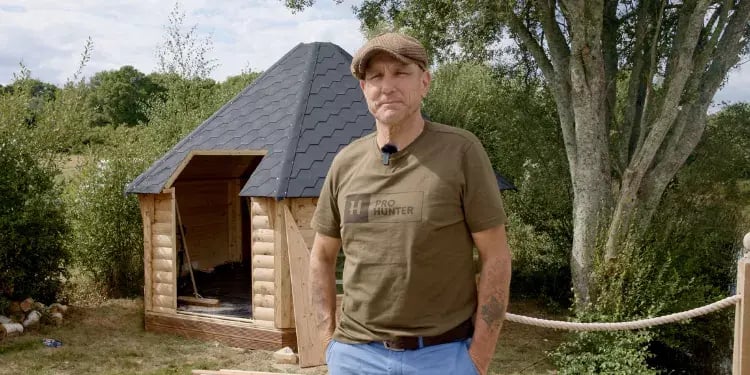 Vinnie Jones stood outside a log cabin that is in the process of being built. Vinnie is wearing blue jeans, a brown t-shirt and a brown tweed flat cap. The log cabin behind him is hexagonal in shape with straight walls and a black roof.