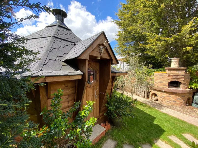 A BBQ hut tucked into the corner of a garden. The cabin is surrounded by trees and shrubs. The is a deer skull with antlers above the cabin door.