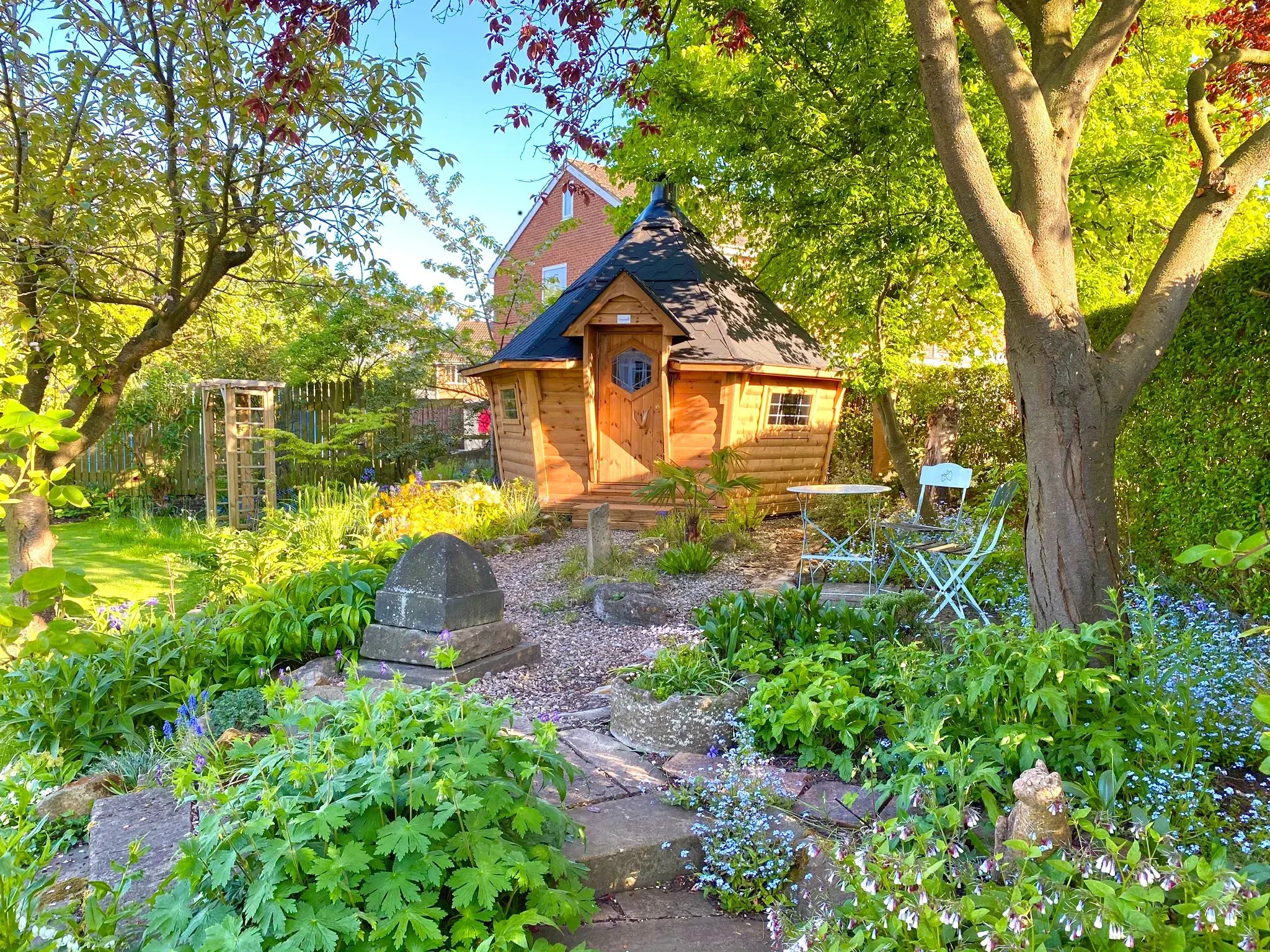 A barbebue hut on a gravel path surrounded by trees, shrubs and flowers.