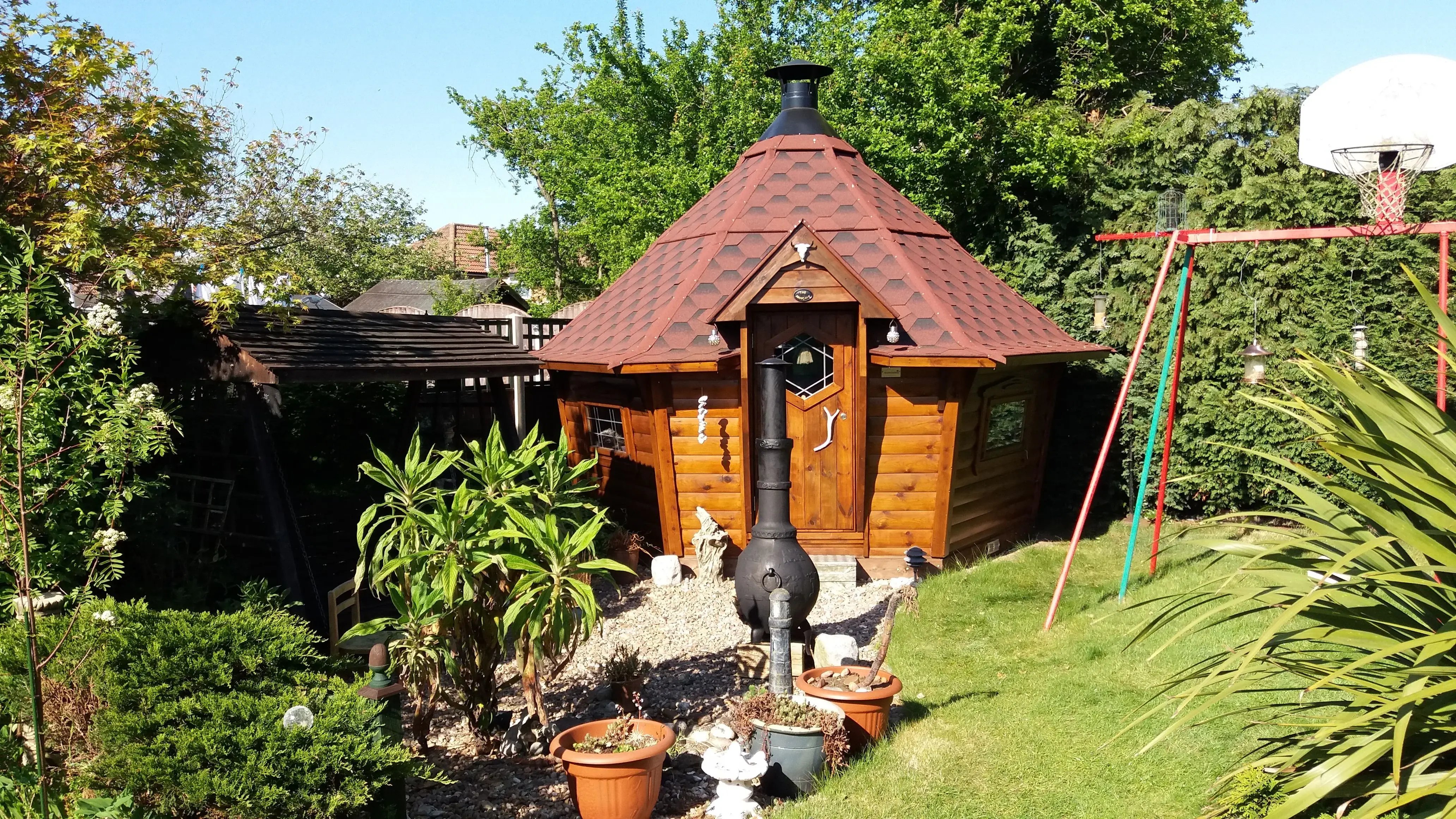A BBQ cabin with a red roof in the corner of a garden.