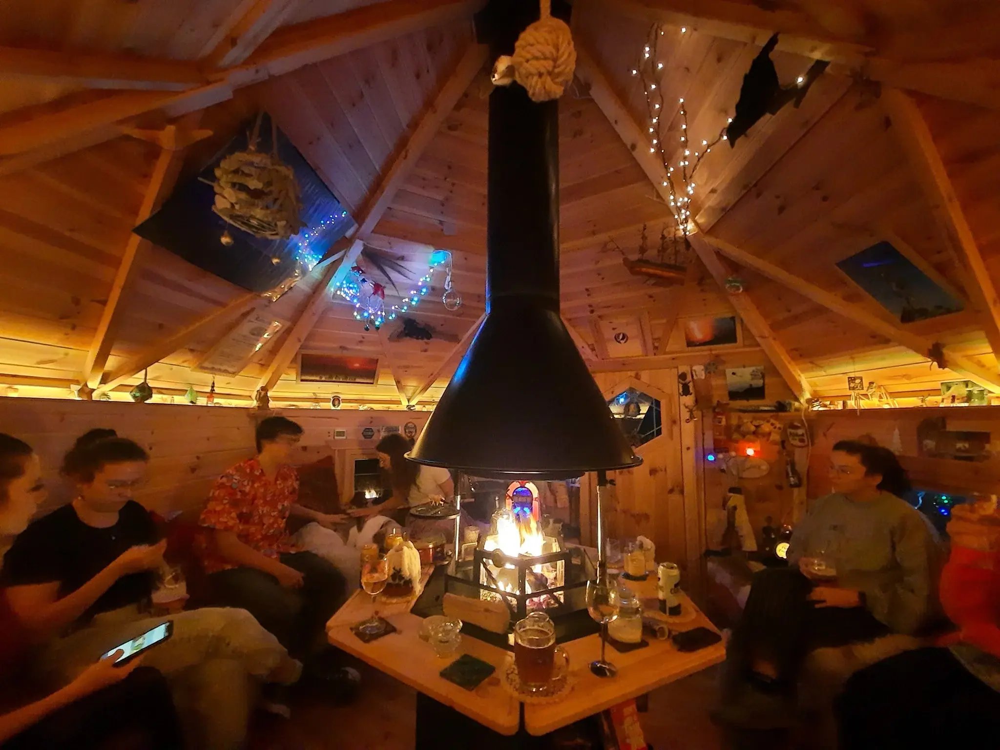 A group of people sat on the benches of a hexagonal BBQ cabin. The cabin is lit warmly and the fire pit has a steady fire lit. On the table around the grill are wine and beer glasses.