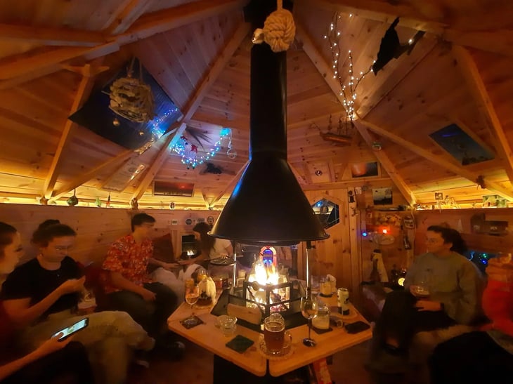 A group of people sat on the benches of a hexagonal BBQ cabin. The cabin is lit warmly and the fire pit has a steady fire lit. On the table around the grill are wine and beer glasses.