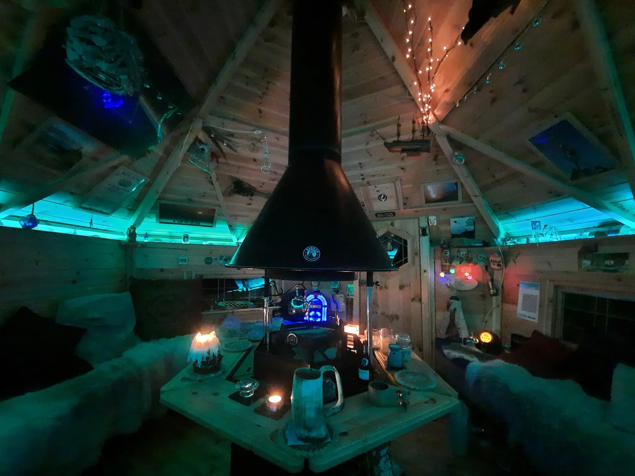 The interior or a barbeque hut, lit in a turquoise light. Around the grill are different sized lit candles and drinking tankards.