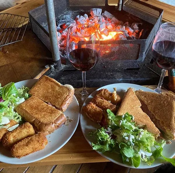 Two plates of toasted sandwiches and salad with glasses of wine in front of the central grill of a barbeque hut.