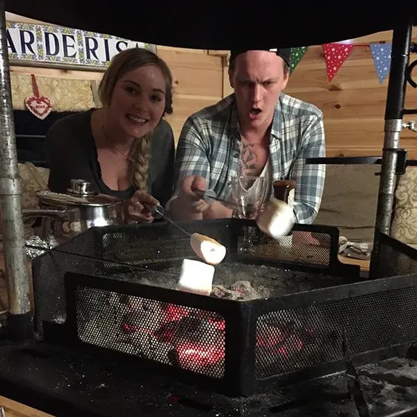Two people sat around a grill in a BBQ hut toasting marshmallows.