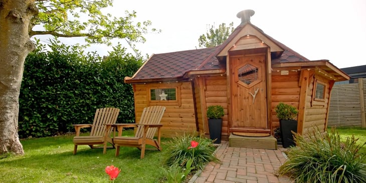 Exterior of a 10m² garden bar with a red roof in a green garden with seating outside and a stone path leading upto the door