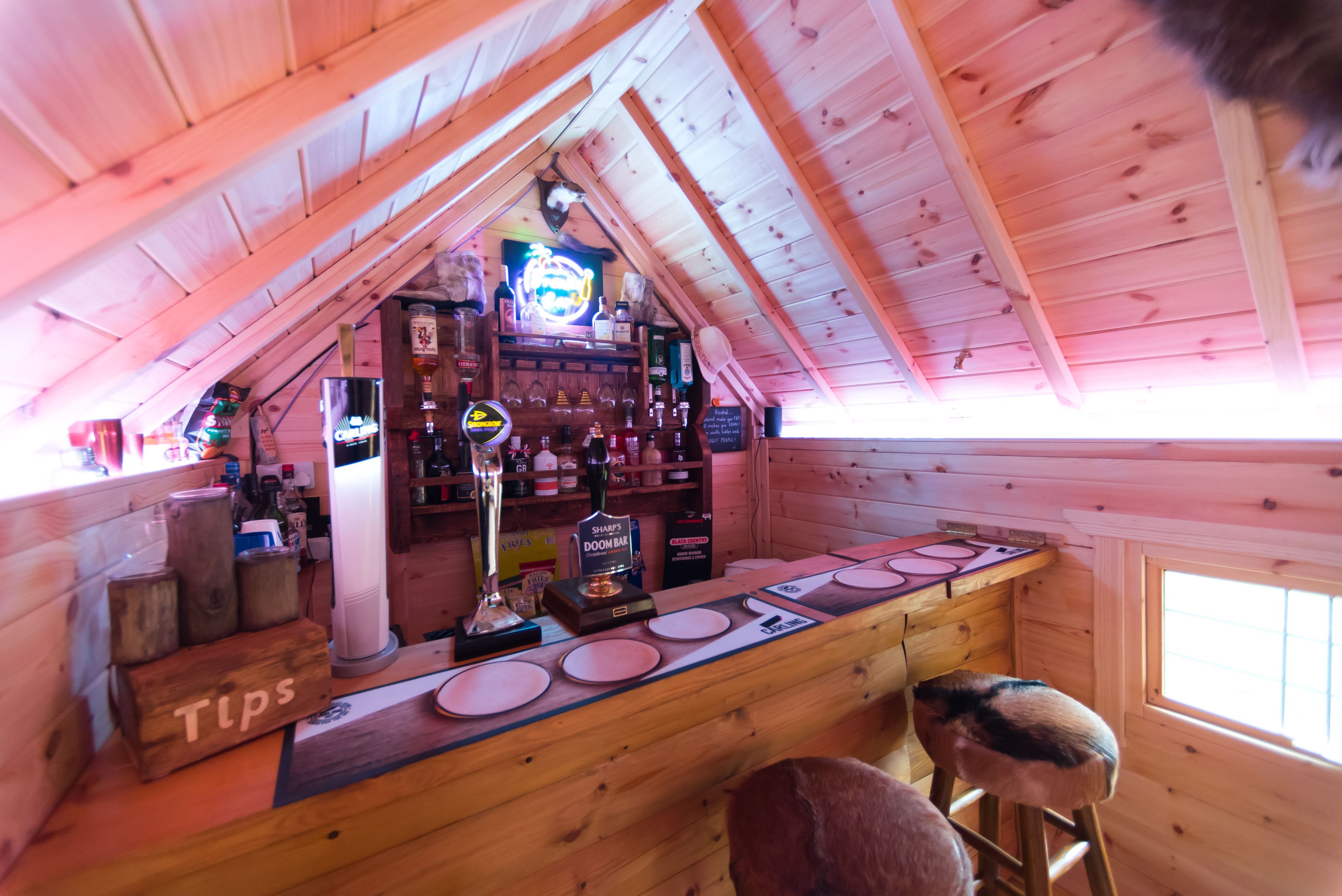 A bar set up in the extension of a BBQ cabin. The are beer pumps on the counter and a drinks rack on the back wall with glasses and bottles of alcohol.