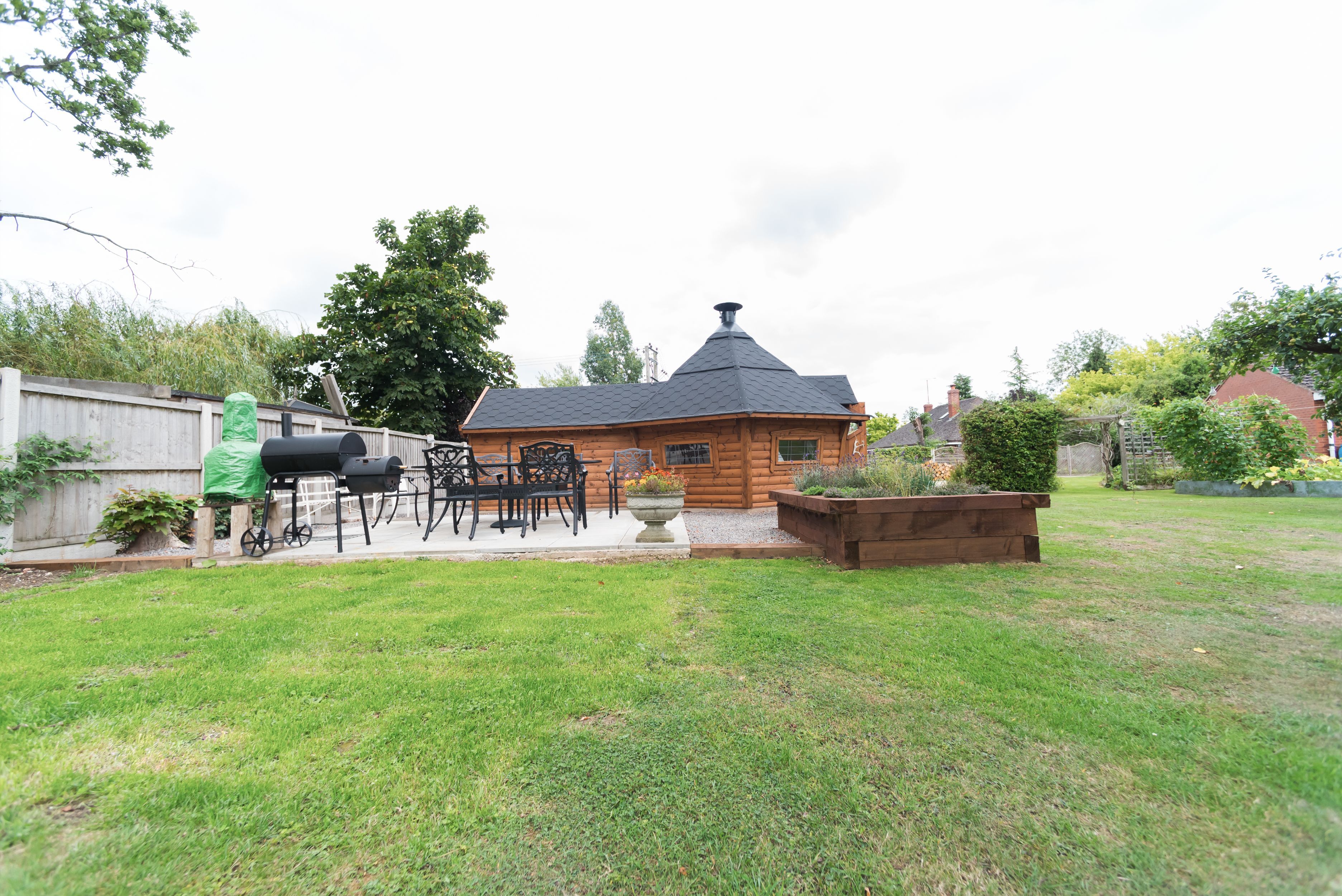 The side view of a BBQ cabin with an extension. In front of the cabin is a patio with an outdoor dining set, smoker and pizza oven.