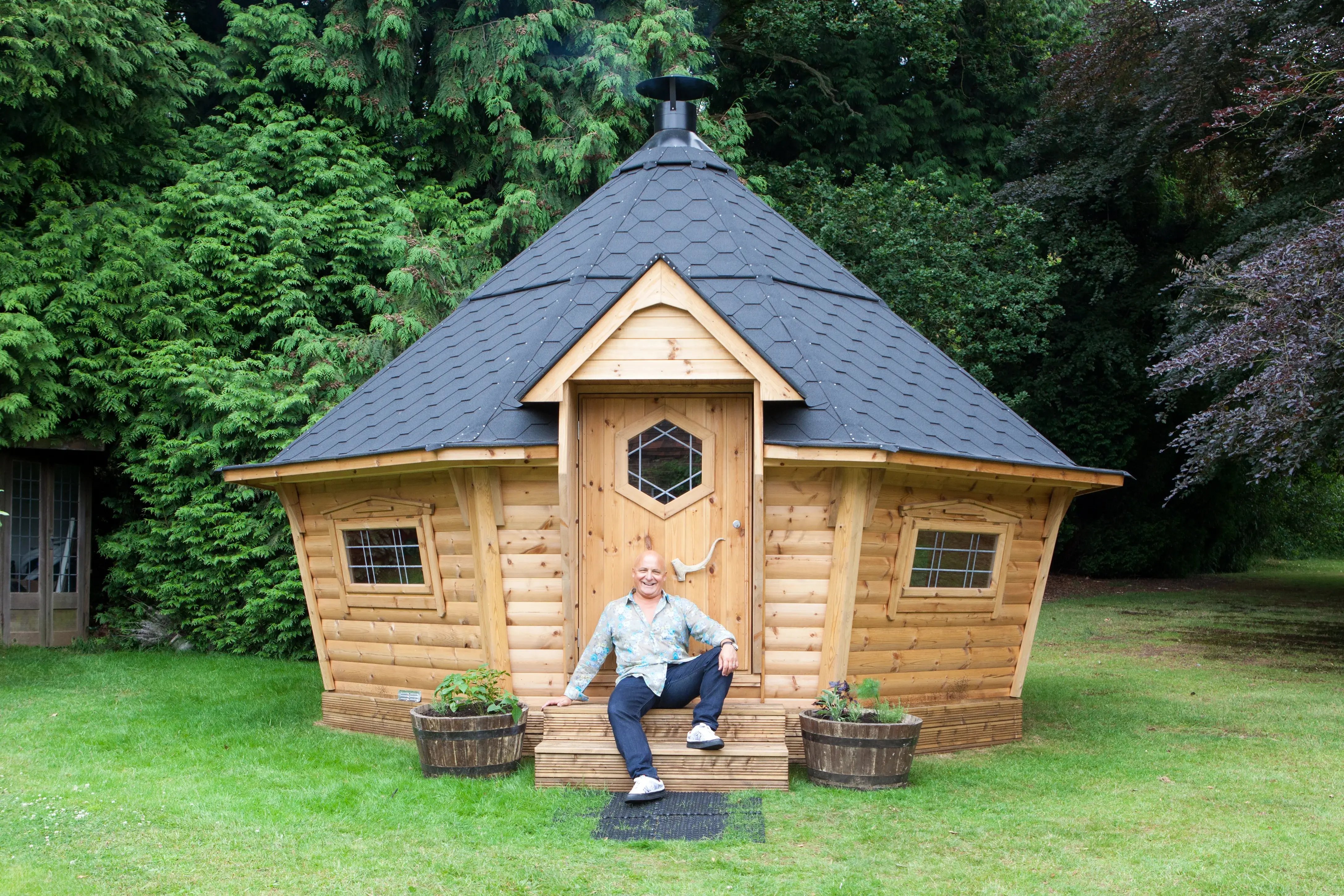 Aldo Zilli sat on the steps of his 17m2 BBQ hut. Aldo is wearing dark blue jeans and a blue & grey floral patterned top. The BBQ hut is on grass and surrounded by trees.
