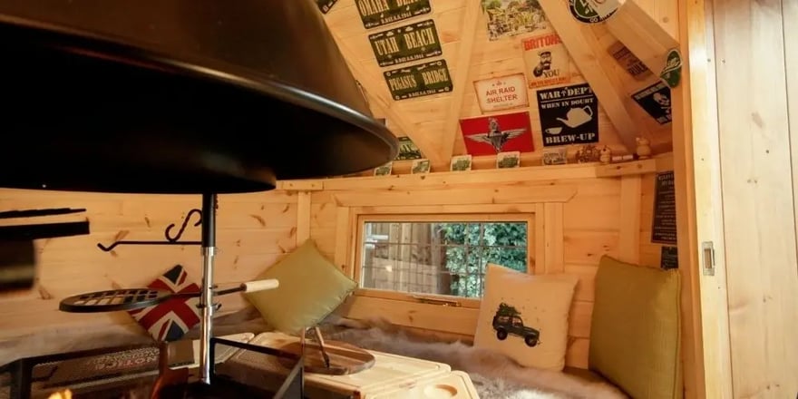 Interior of a small BBQ hut and wooden grill cabin with wraparound seating, cushions, a central barbecue and Land Rover signs.