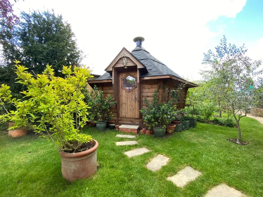 A 10m2 BBQ hut with a black roof, surrounded by trees and shrubs with a rectandular stone path leading to the door.