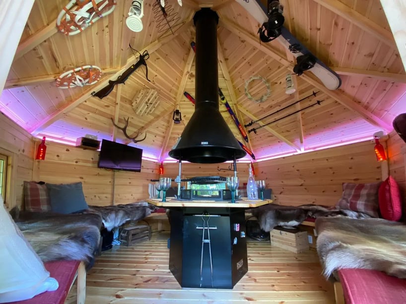 Inside a hexagonal log cabin with a central BBQ grill. The roof of the cabin is decorated with rustic skiing a snow sport equipment.