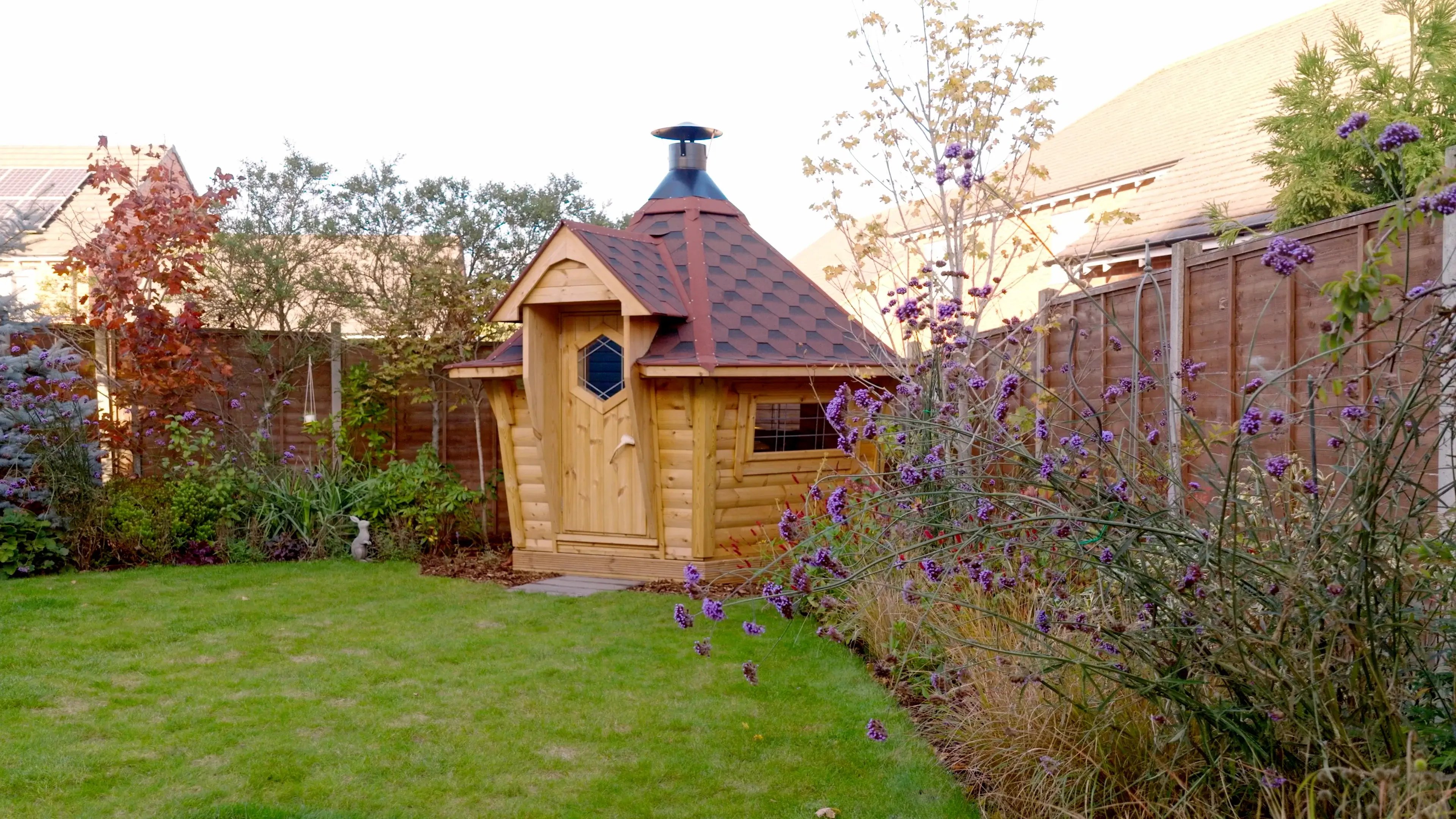 A 7m2 BBQ cabin with a red roof in the corner of a garden.