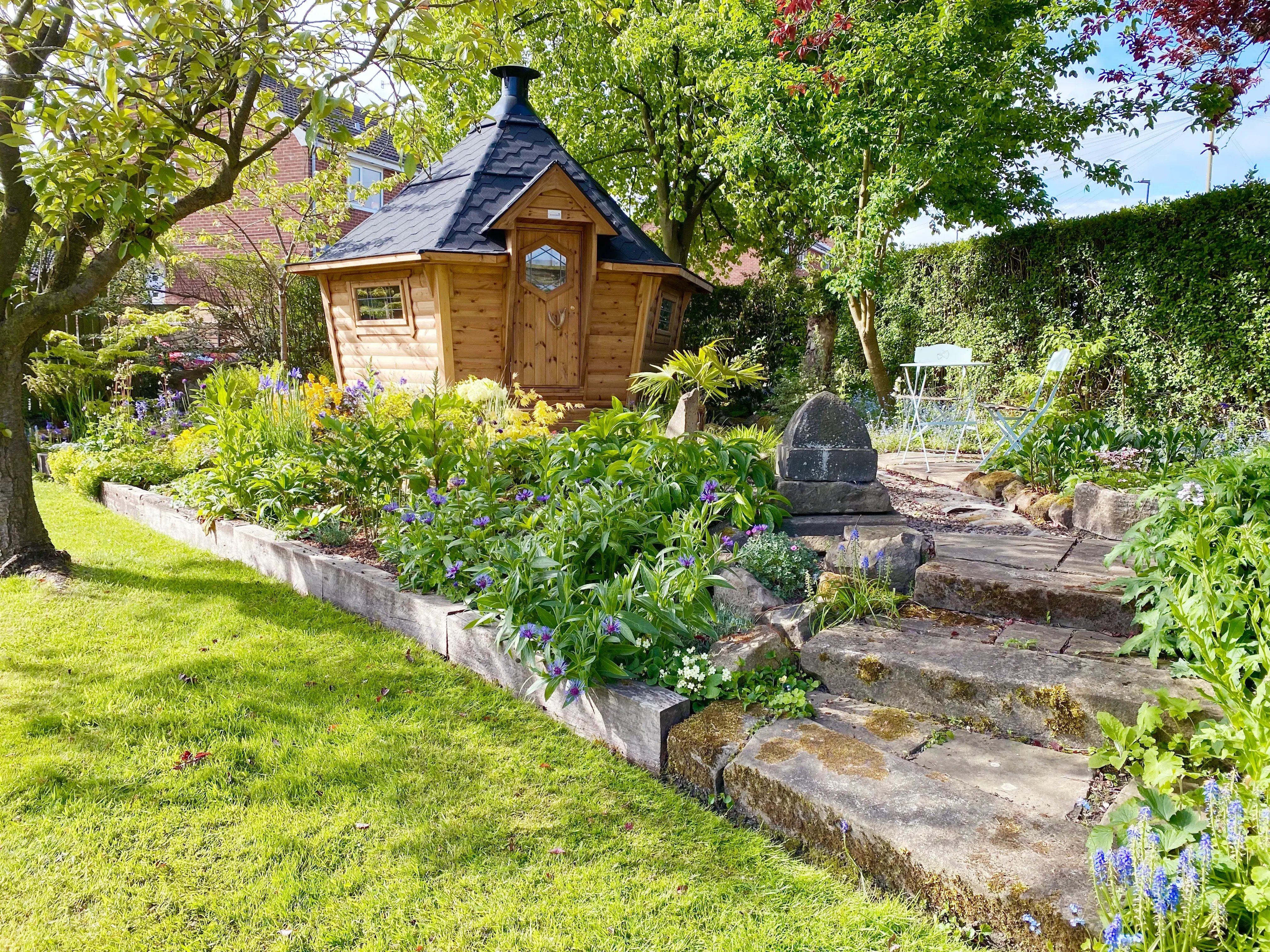 A hexagonal barbeque hut sat on a raised platform surrounded by trees, shrubs and flowers. Ther are stone steps leading up to the cabin and a metal table and chairs at the front.