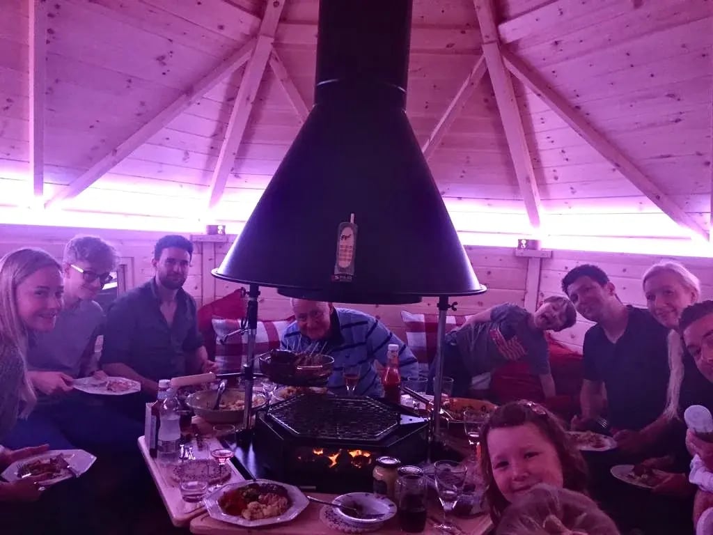 People huddled around a indoor BBQ grill. Food is peing cooked on the barbeque while people sit around the cabin.