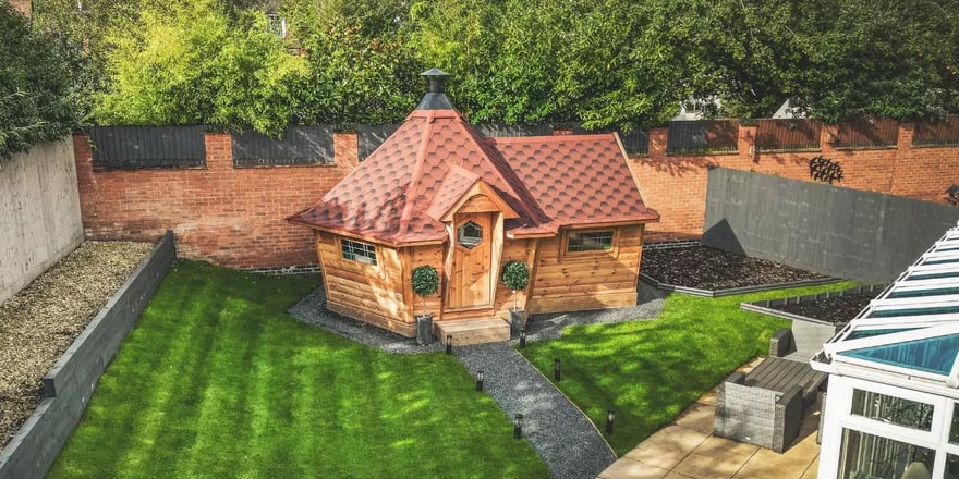 A BBQ hut with a side extension and red roof at the bottom of a garden.