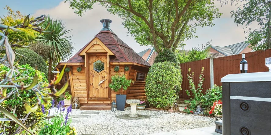 A 10m2 BBQ hut with a red roof in a garden surrounded by bushes and grey gravel.