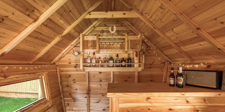 The bar area in an Arctic Cabins garden bar. There are various drinks and glasses on the drinks rack with fairy lights lining the edge of the cabin. On the bar there are some beers and a radio.