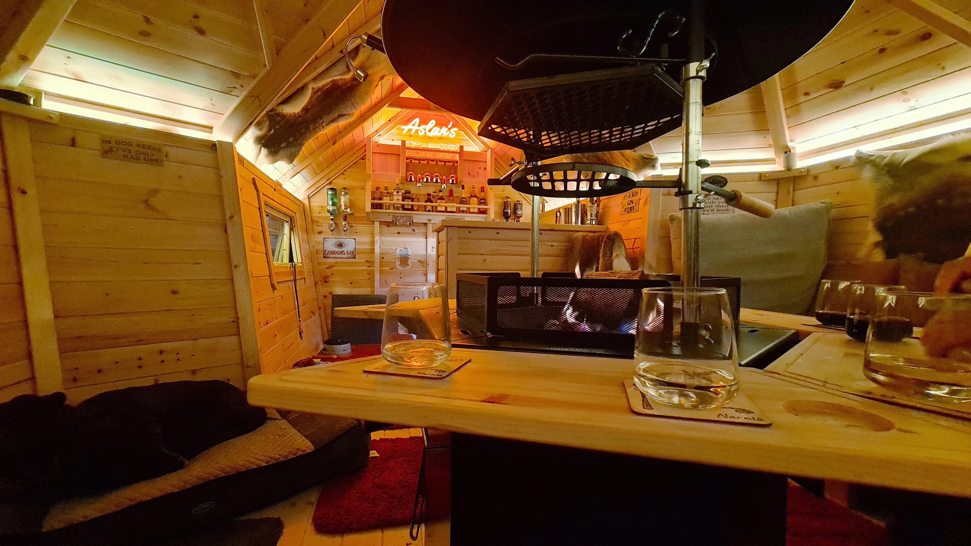 A warmly lit garden bar with drinks on the table and a small fire in the grill.