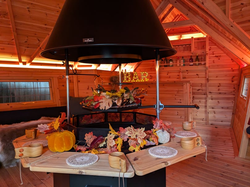 The interior of a log cabin bar, lit with soft orange lighting. The central grill is decorated with faux a leaf garland and pumpkins. On the counter there are small LED letters spelling "BAR" and a drinks rack with bottle on the far wall.