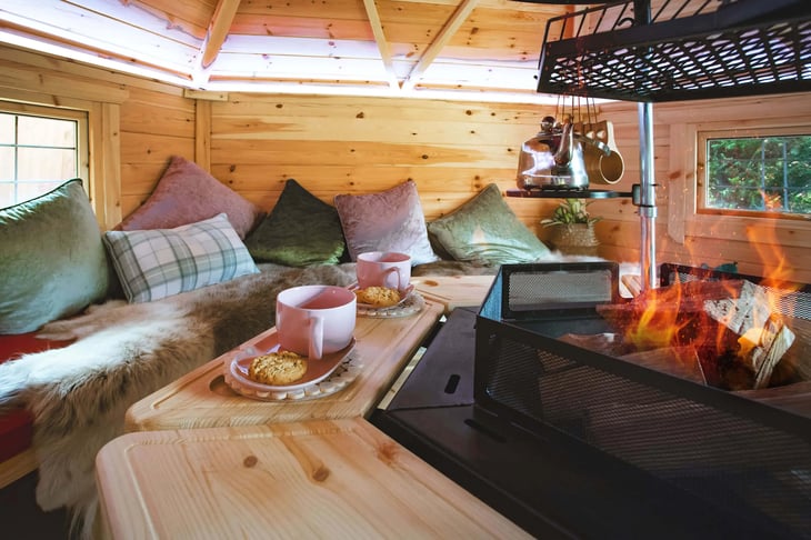 Two pink mugs on plates with chocolate chip cookies sat on the table that surrounds the central gill in a BBQ cabin. The benches around the cabin walls are decorated with reindeer skin throws and pink & green cushions.