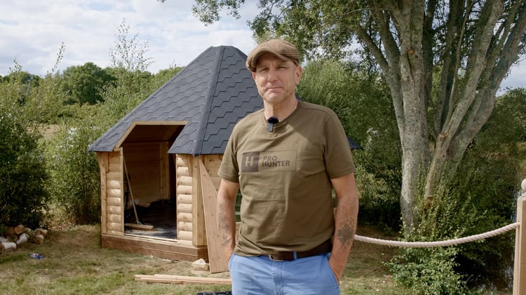Vinnie Jones stood outside a log cabin that is in the prcoess of being built. Vinnie is wearing blue jeans, a brown t-shirt and a brown tweed flat cap. The log cabin behind him is hexagonal in shape with straight walls and a black roof.