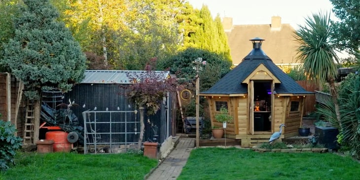 Small Scandinavian BBQ hut in a garden, showing the wooden outdoor hut, pitched roof, chimney and open cabin doorway.