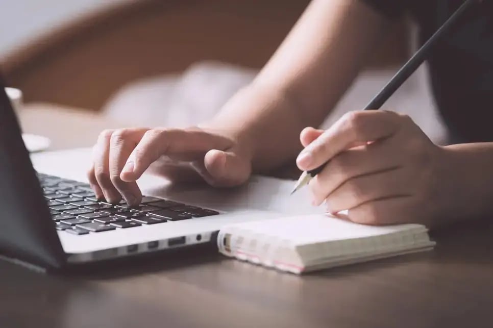 person writing in notepad next to laptop