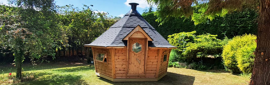 10m BBQ Cabin by Arctic Cabins, on grass with tree to left, BBQ grill, black pointy roof