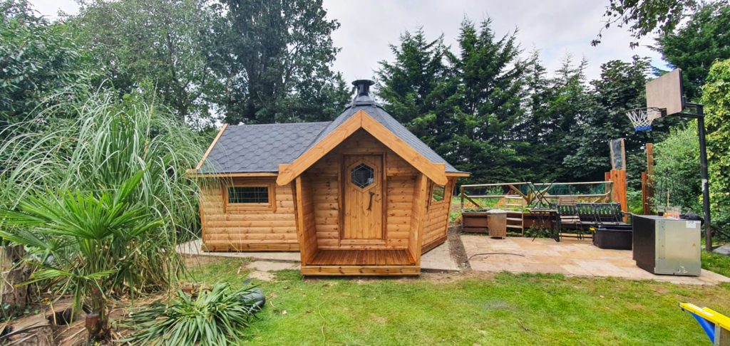 10m Arctic Bar by Arctic Cabins, black roof, in garden with grass and basket ball net to the right of the image. BBQ grill unit as well as a Bar extension. Contact Us